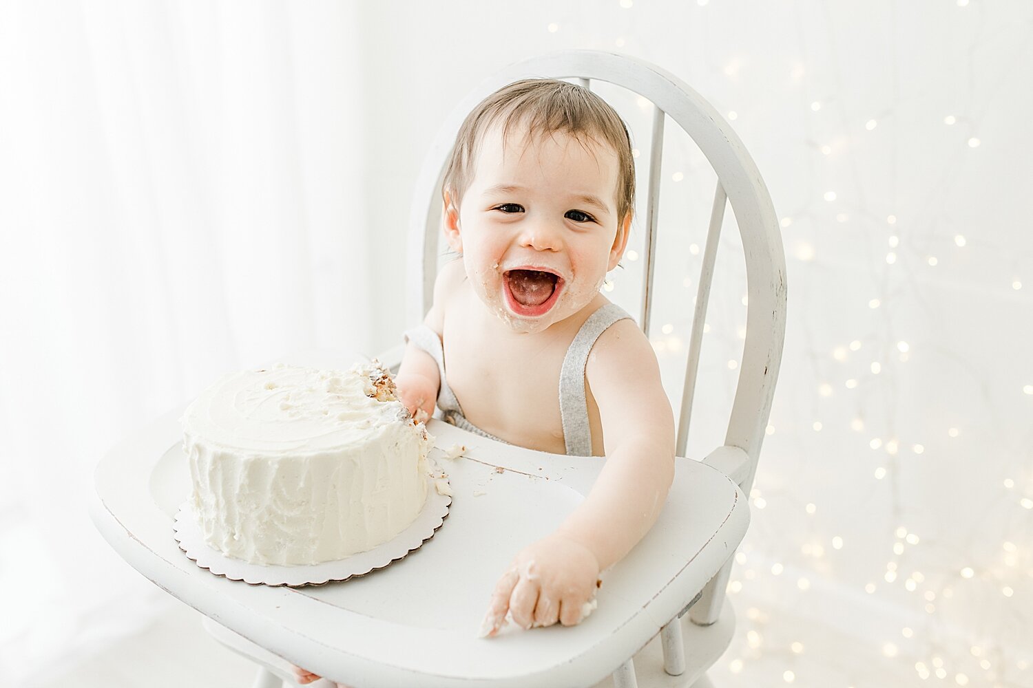 First birthday cake smash for a little boy. Twinkle lights are hanging behind him. Photos by Darien Photographer, Kristin Wood Photography.