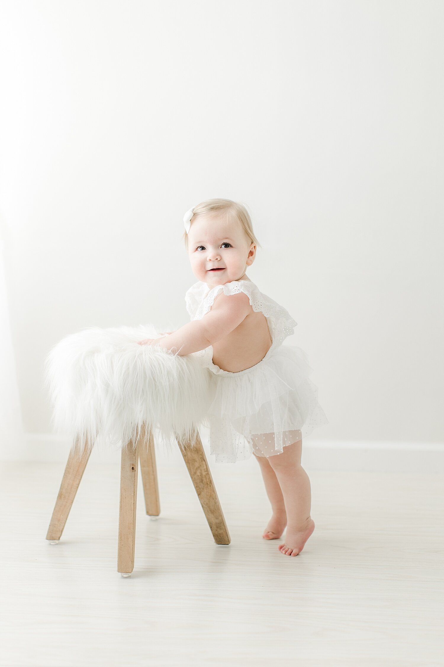 Baby girl in all white standing by a stool for photos for her first birthday. Photo by Kristin Wood Photography.