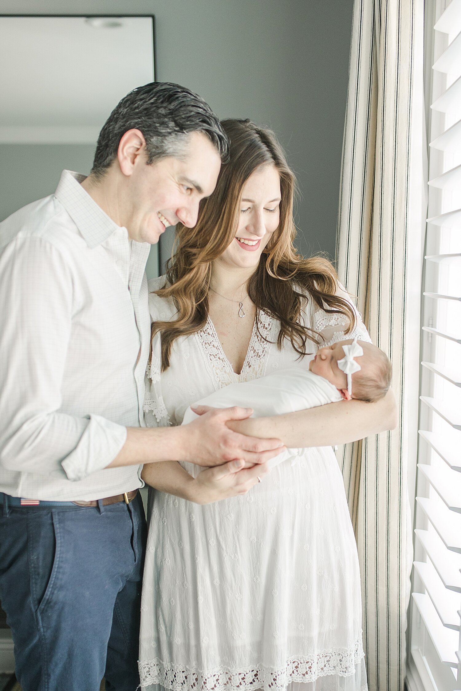Mom and Dad standing by window with newborn. Photo by Kristin Wood Photography.