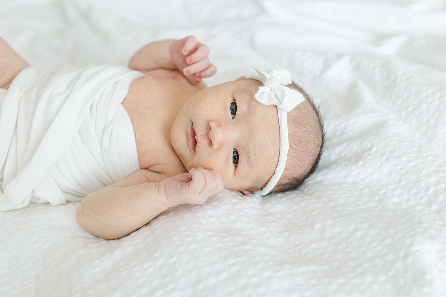 Photo of baby girl laying on a bed in all white wide awake. Photo by Kristin Wood Photography.