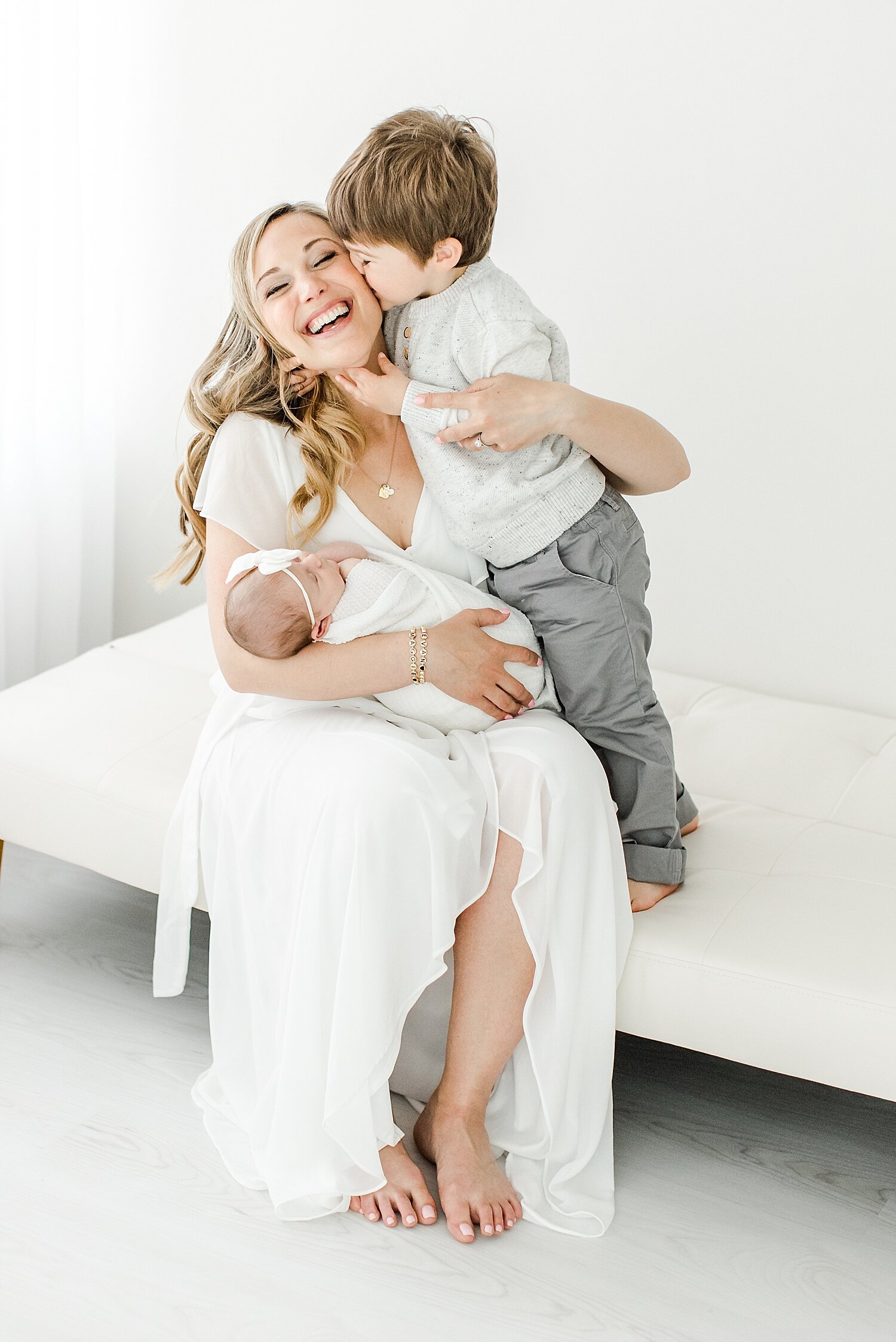 Toddler giving Mom a hug and kiss while she holds his baby sister. Kristin Wood Photography is sharing tips for how to have a successful newborn session with a toddler.
