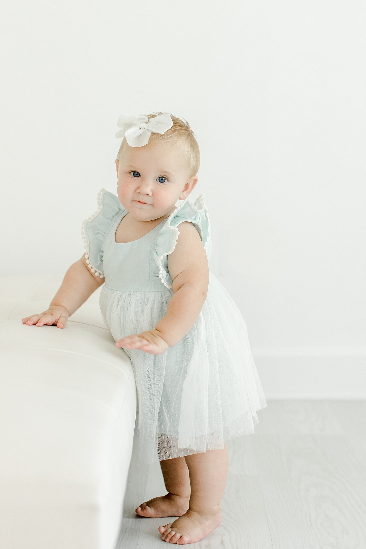 8 month old baby girl standing by couch in studio for photos with Kristin Wood Photography.