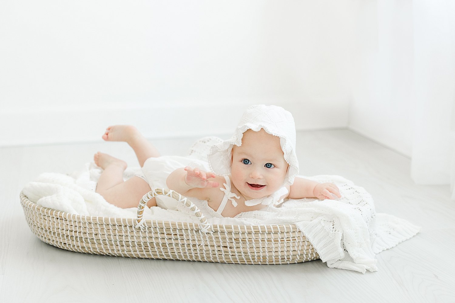 8 month old baby girl wearing a white bonnet, sitting in a Moses basket | Kristin Wood Photography