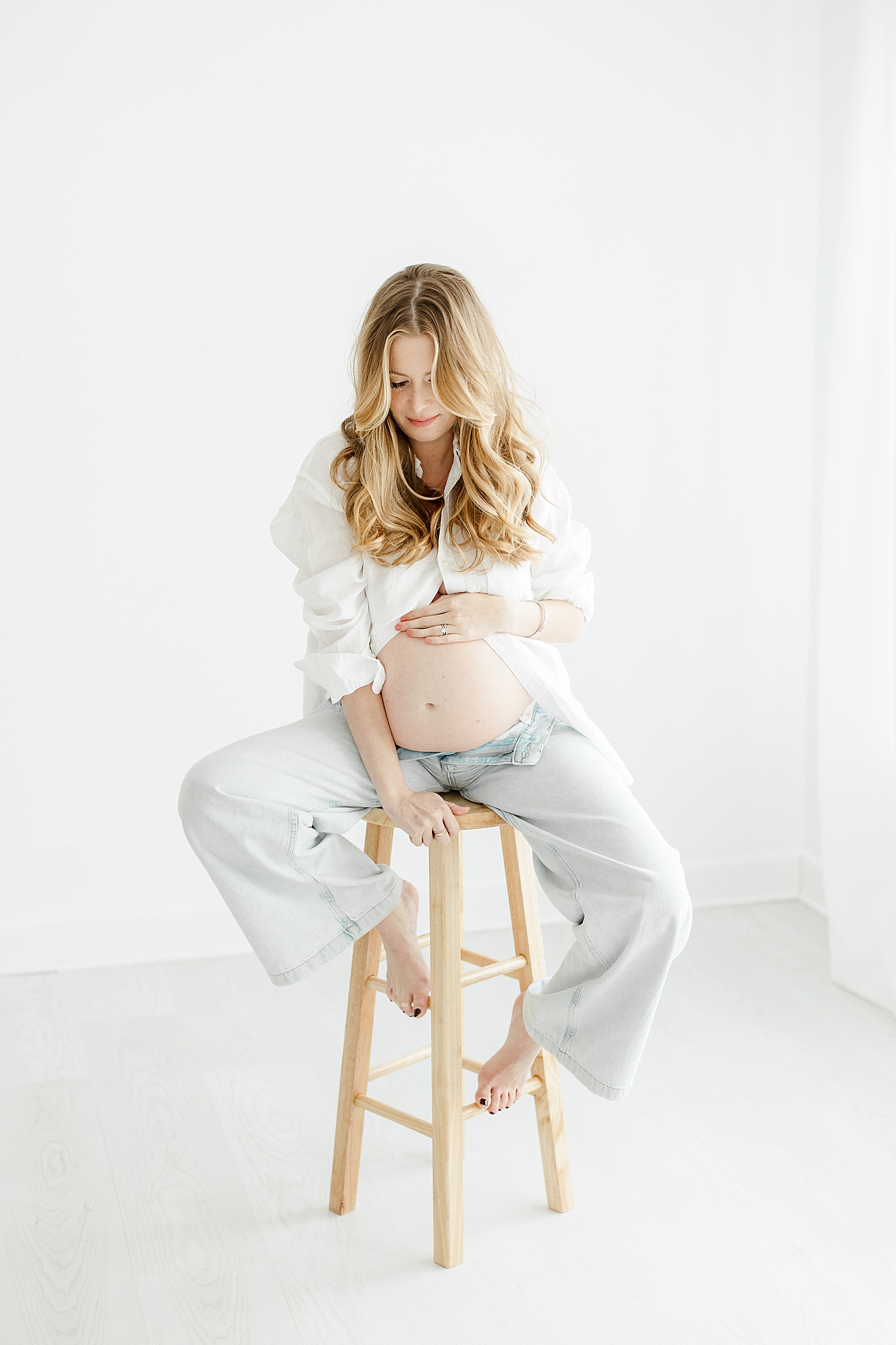 What To Wear for Your Maternity Session | Expecting mom sitting on stool wearing denim and a white button-up shirt showing pregnant belly | Kristin Wood Photography