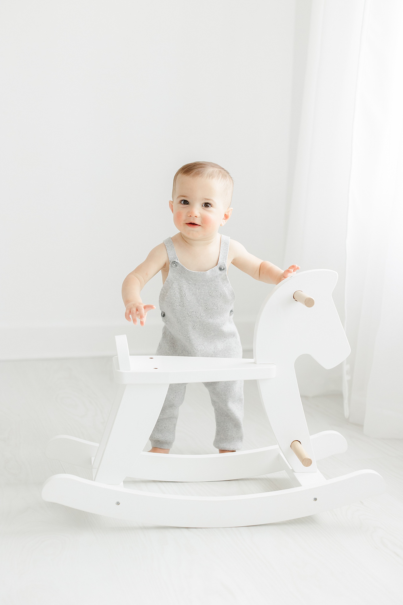 One year old boy standing with rocking horse during first birthday photoshoot with Kristin Wood Photography.